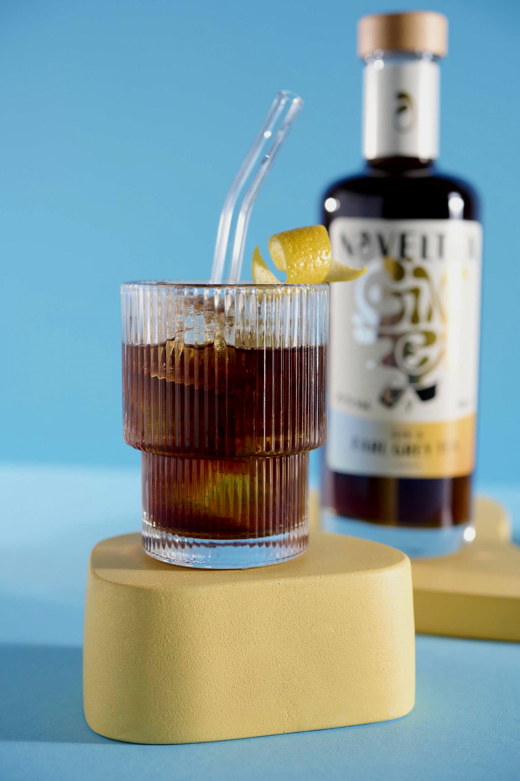 Noveltea, Gin & Tea mit Longdrink auf gelbem Sockel vor blauem Grund